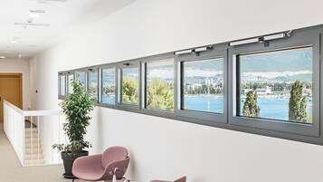 A modern interior with a row of windows offering a view of a body of water and greenery. There are two chairs and a small table in the foreground.