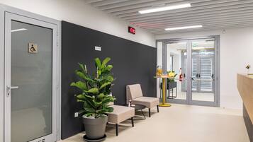 Un espace d’attente moderne avec une porte en verre, une plante en pot, deux chaises, une petite table et une horloge numérique.