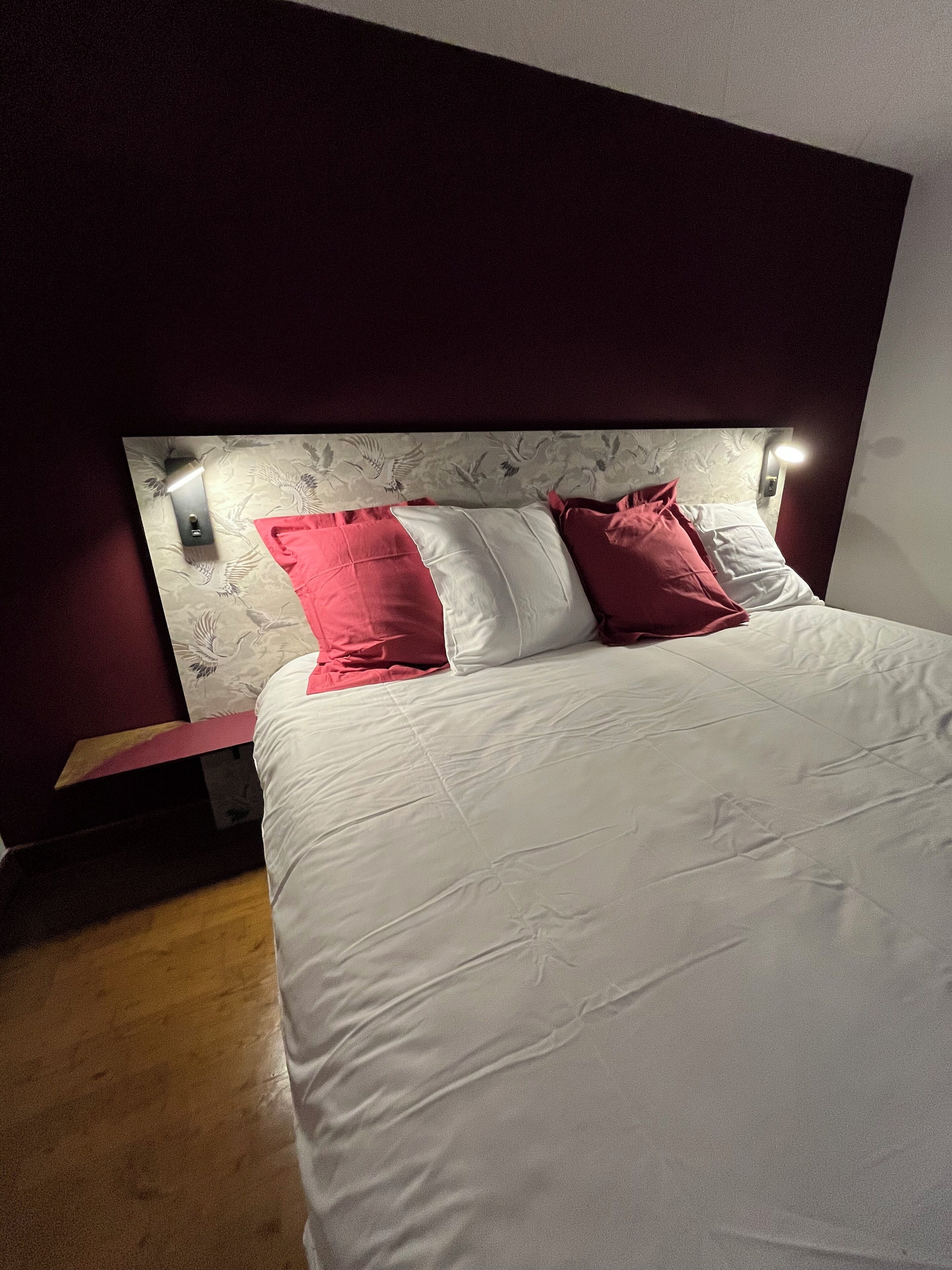 Un lit bien fait, avec une literie blanche et rouge, une tête de lit ornée de motifs floraux et un luminaire fixé au mur.