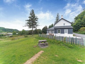 A house with a white exterior and dark trim, a fenced yard, a picnic table, and a view of a river and hills in the background. - The location sits beside the Atlantic bridge, offering uninterrupted sea views. (Clachan Seil)