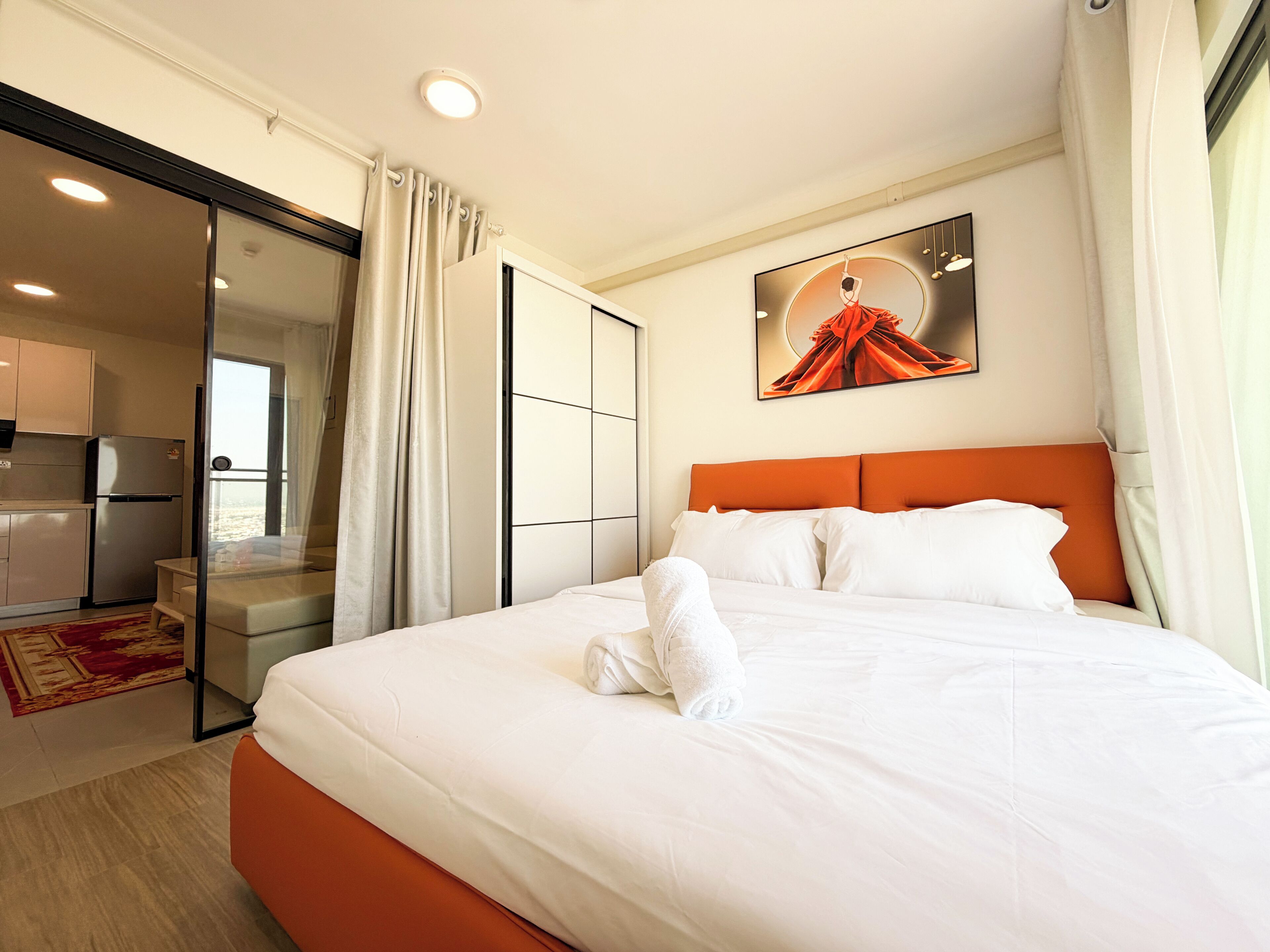 A hotel room with a bed, a painting, a sliding door to a kitchen, and a view of the outside.