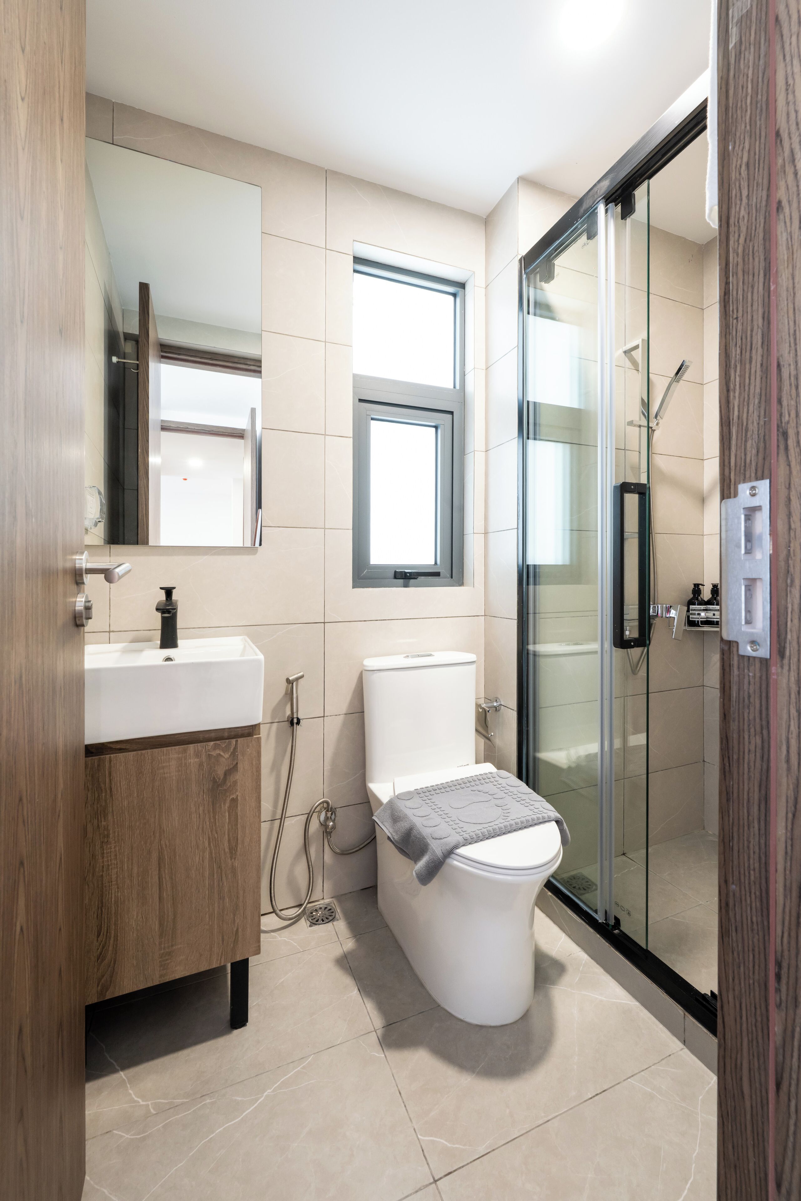 A modern bathroom with a wooden vanity, a toilet, a glass shower enclosure, and a window.