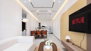 Una sala de estar moderna con un sofá blanco, una mesa de centro de madera con un control remoto, un televisor montado en la pared y un comedor al fondo.