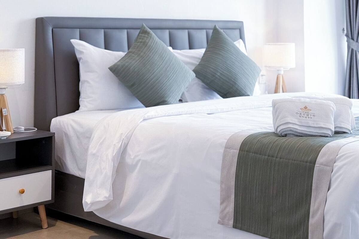 Una cama bien arreglada con sábanas blancas y dos almohadas grises, una mesita de noche con una lámpara y un cabecero gris.
