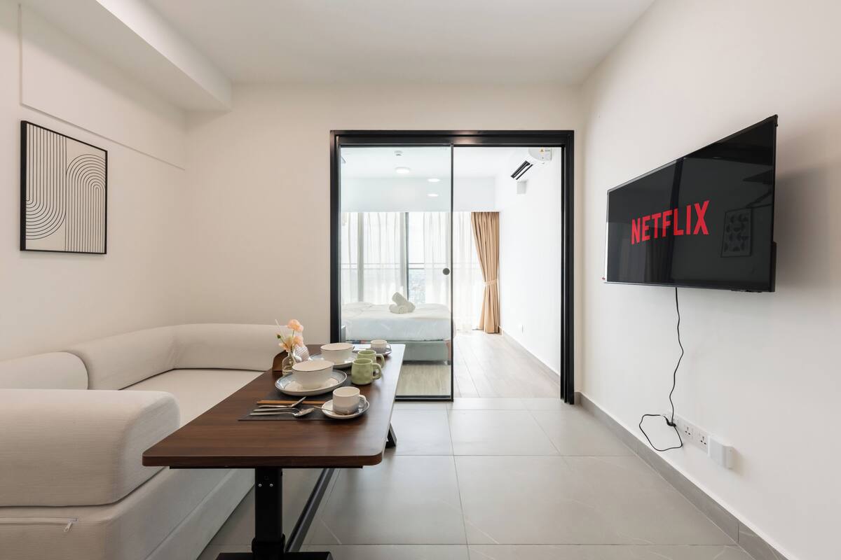 Una habitación de hotel moderna con comedor, sofá, televisor montado en la pared y puerta corrediza de vidrio que da a un balcón.
