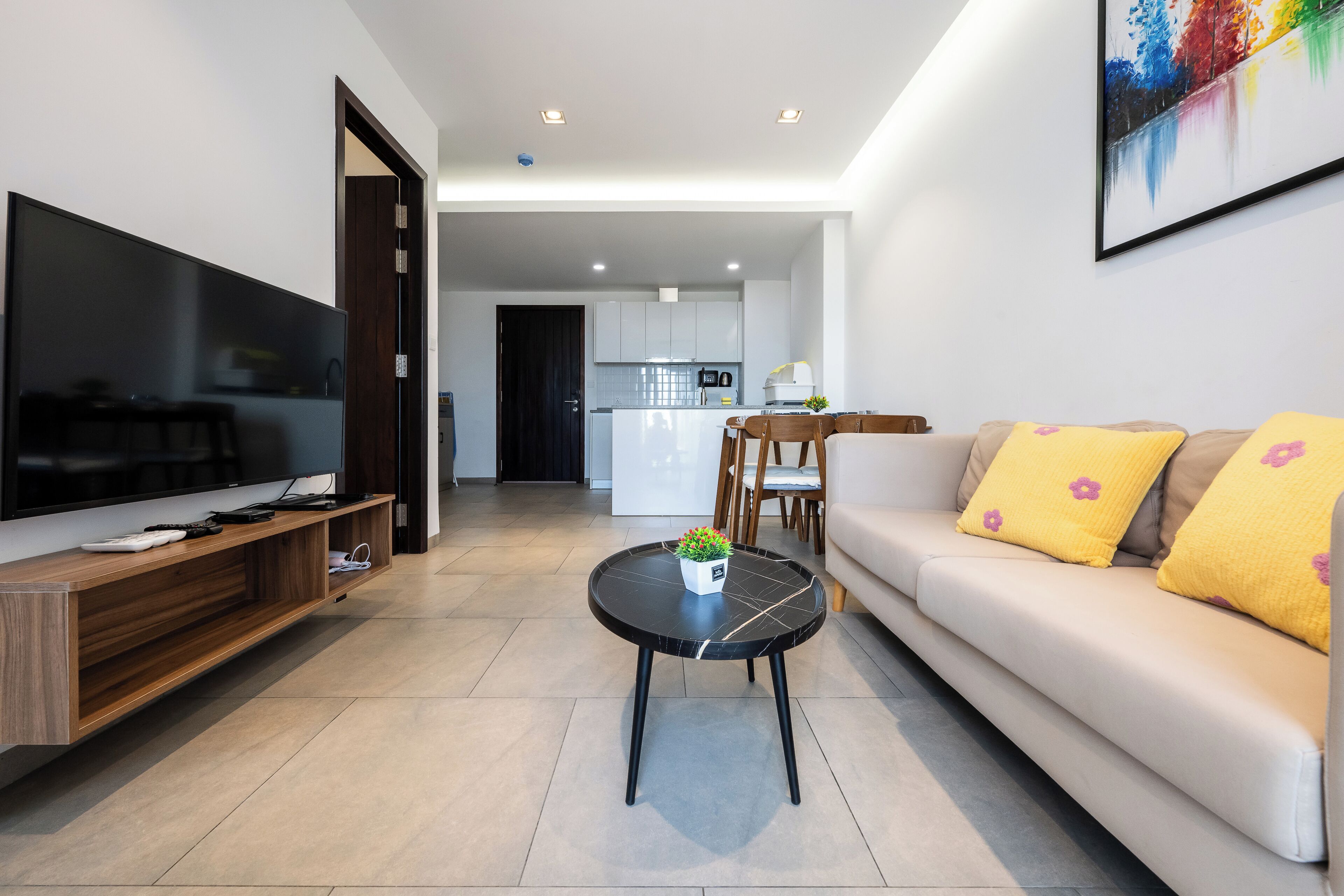 A modern living room with a flat-screen TV, wooden entertainment unit, a round coffee table with a plant, a beige sofa with yellow pillows, and a kitchen in the background.