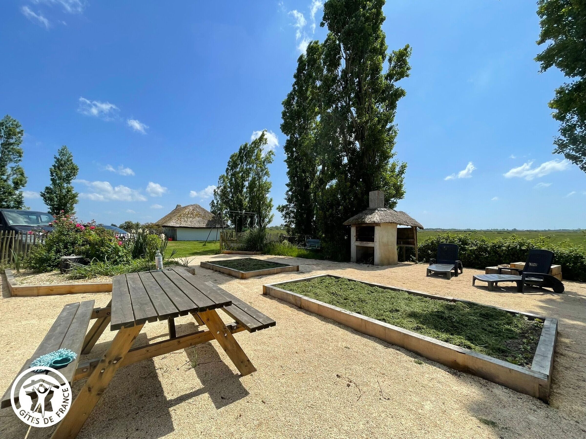 Un cadre extérieur rustique avec une table de pique-nique en bois, une petite structure à toit de chaume et un espace jardin avec des plates-bandes surélevées.