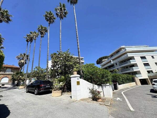 Un espace de stationnement avec une voiture noire, des palmiers et un bâtiment blanc doté de nombreux balcons.