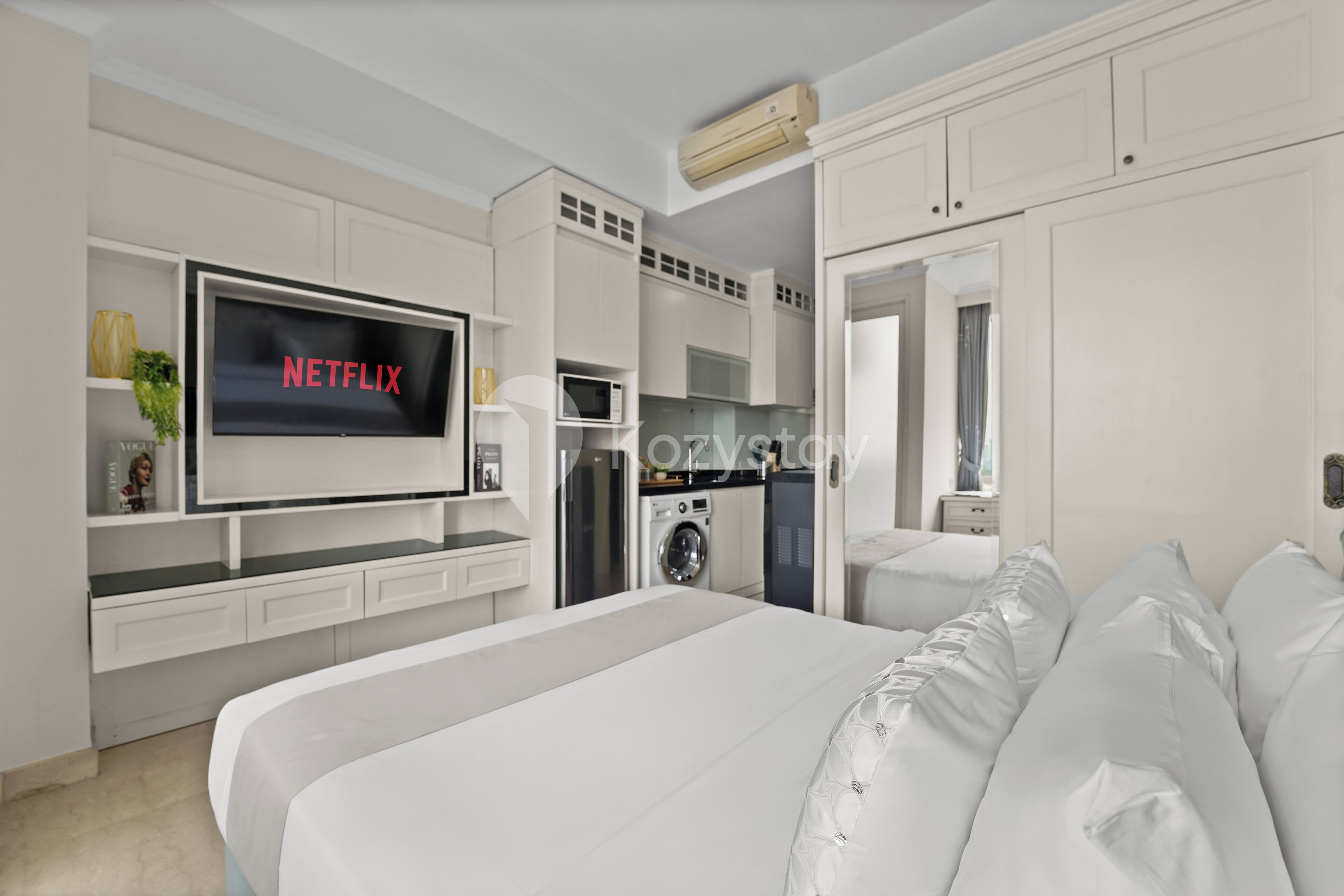 A modern bedroom with a large bed, a TV mounted on the wall, a built-in washing machine, and a kitchenette.