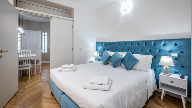 Ein Schlafzimmer mit einem großen Bett, einem blauen, gepolsterten Kopfteil, weißer Bettwäsche, einem Nachttisch mit Lampe und Blick auf einen Essbereich durch eine offene Tür.