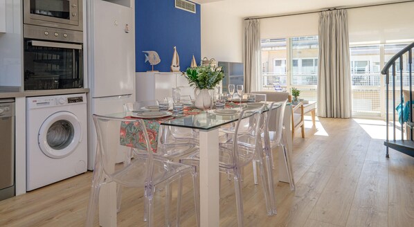 Une cuisine moderne dotée d’appareils électroménagers en acier inoxydable, une table à manger en verre avec des chaises transparentes, et une vue sur la ville grâce à de grandes fenêtres.