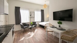 Ett modernt, väl upplyst rum med ett litet kök, en matplats med bord och stolar, en säng med gardiner och en tv som är monterad på väggen.