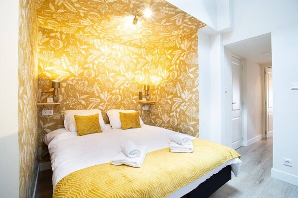 A bedroom with a floral wallpaper, a bed with white and yellow bedding, and a bedside table with a lamp.