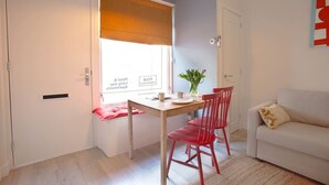 Een kleine kamer met een eettafel, twee rode stoelen, een witte bank en een raam met een zonnescherm.