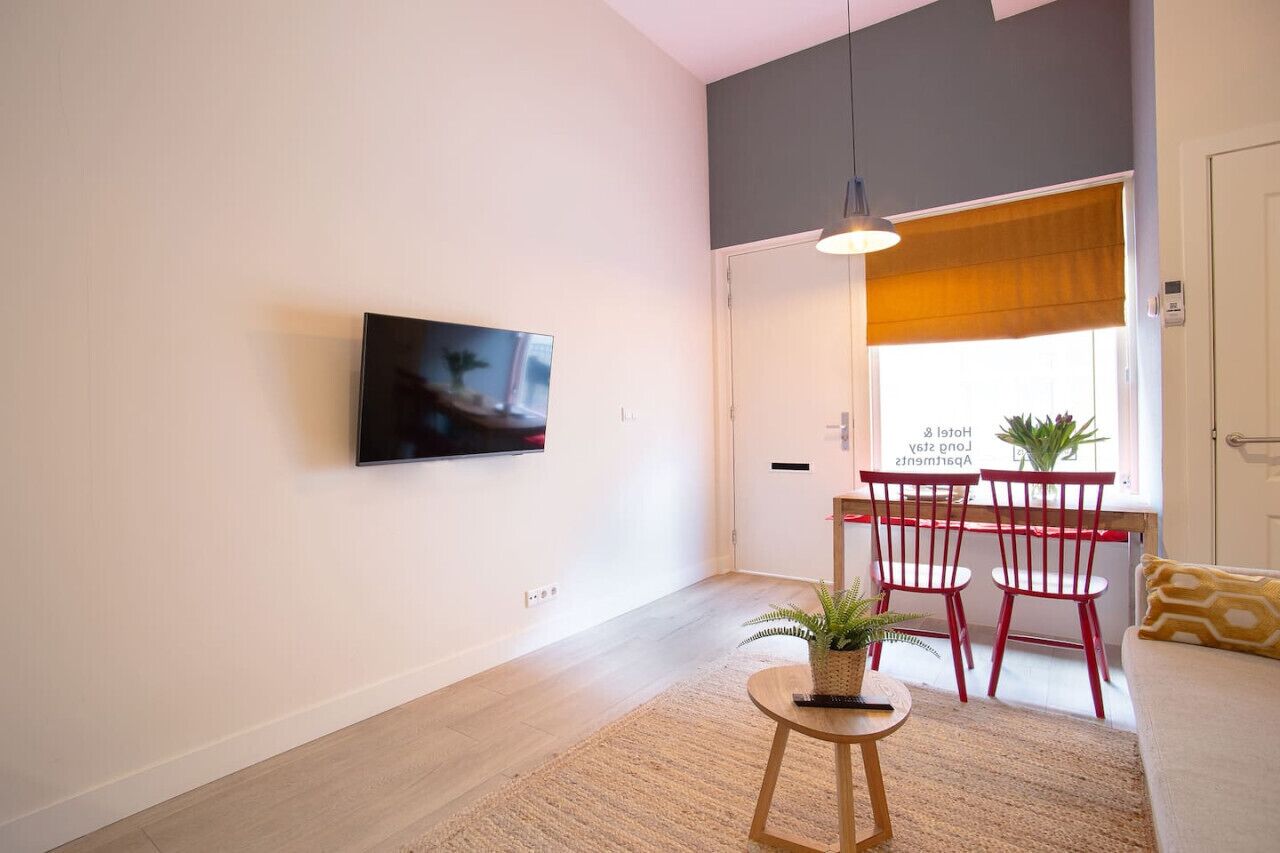 En moderne stue med et fladskærms-tv på væggen, et rundt træbord med en plante, røde stole og en sofa med en gul pude.