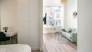 A modern, well-lit room with a bed, a desk, a sofa, and a dining area with a view of buildings through large windows. - Modern apartment near Scheveningen beach (Den Haag)