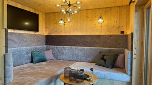 A cozy room with a wooden ceiling, a large flat-screen TV mounted on the wall, a modern chandelier, and a cushioned seating area with a small table. - Panoramic hut in the ski area (Wildschönau-Niederau)