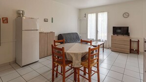 A compact kitchen with a refrigerator, a dining table with chairs, a sofa, a television, and a clock on the wall.