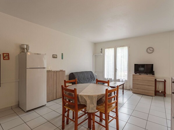 A compact kitchen with a refrigerator, a dining table with chairs, a sofa, a television, and a clock on the wall.