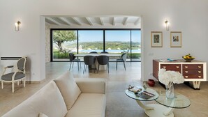 Amplia sala de estar con un ventanal que ofrece vistas a una piscina y jardines. Hay un sofá, una mesa de centro de vidrio y un mueble de madera con cajones.