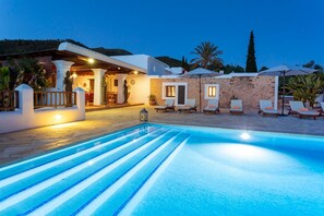 Zona junto a la piscina con sillones reclinables, un muro de piedra y un edificio con terraza cubierta.