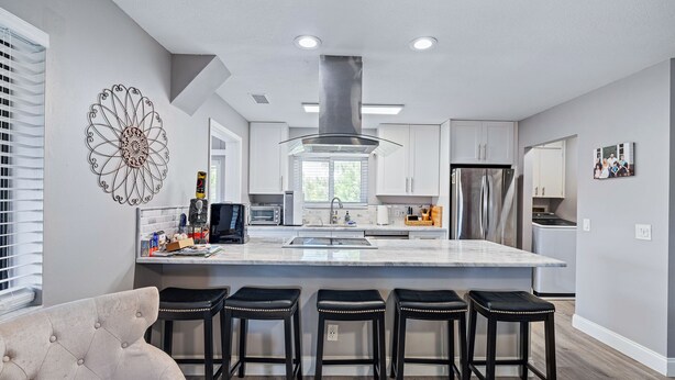 A modern kitchen with a central island, bar stools, and a decorative wall piece. - Spacious 3BR, 2BA beach home just a short walk from the sand! Three bedrooms up, private queen room and bath down. Secure 2-car garage for vehicles or a small boat. Perfect for families or groups. 30-day minimum stay required. (New Smyrna Beach)