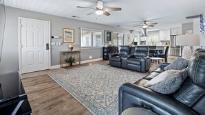 A spacious living room with a large black leather sofa, a matching armchair, and a dining area with a table and chairs. There are ceiling fans, a kitchen in the background, and a door with a white frame. - Spacious 3BR, 2BA beach home just a short walk from the sand! Three bedrooms up, private queen room and bath down. Secure 2-car garage for vehicles or a small boat. Perfect for families or groups. 30-day minimum stay required. (New Smyrna Beach)