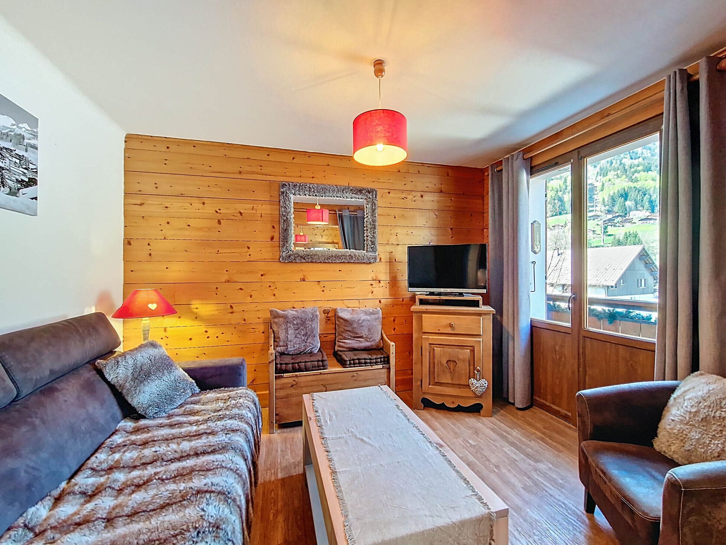Una habitación acogedora con paredes de madera, un sofá, una mesita, un televisor y una ventana con vistas a casas.