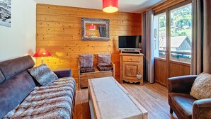 A cozy room with wooden walls, a sofa, a small table, a TV, and a window with a view of houses. - La Clusaz - Apartment 6 ppl, 53 sqm, 2 bedrooms, WiFi (La Clusaz)