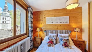 A bedroom with a wooden wall, a bed with white bedding, two bedside tables with lamps, and a window with a view of a building and trees. - La Clusaz - Apartment 6 ppl, 53 sqm, 2 bedrooms, WiFi (La Clusaz)