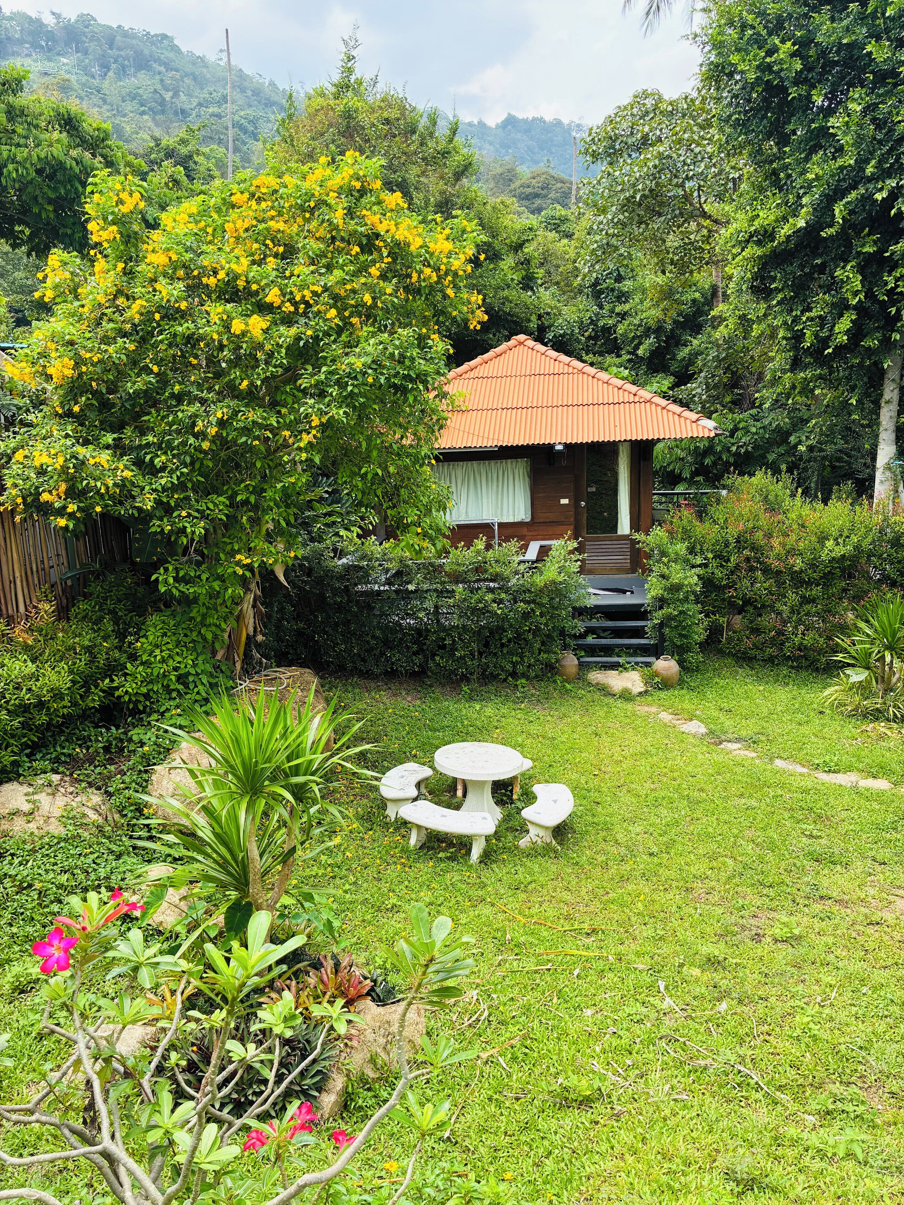 A small house with a red roof, surrounded by lush greenery and a garden with a table and benches.