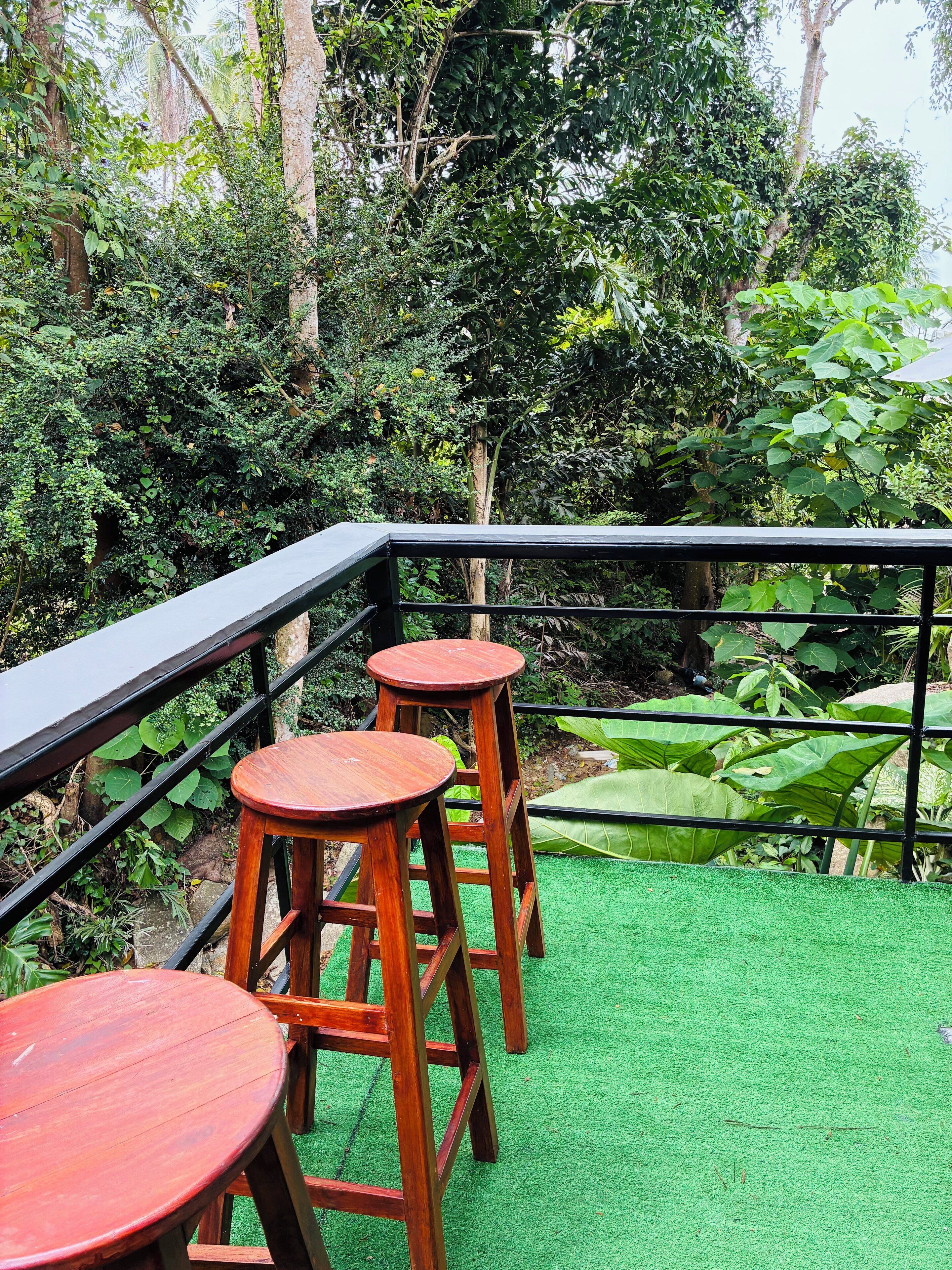 A balcony with wooden stools and a green artificial turf floor, surrounded by dense foliage.