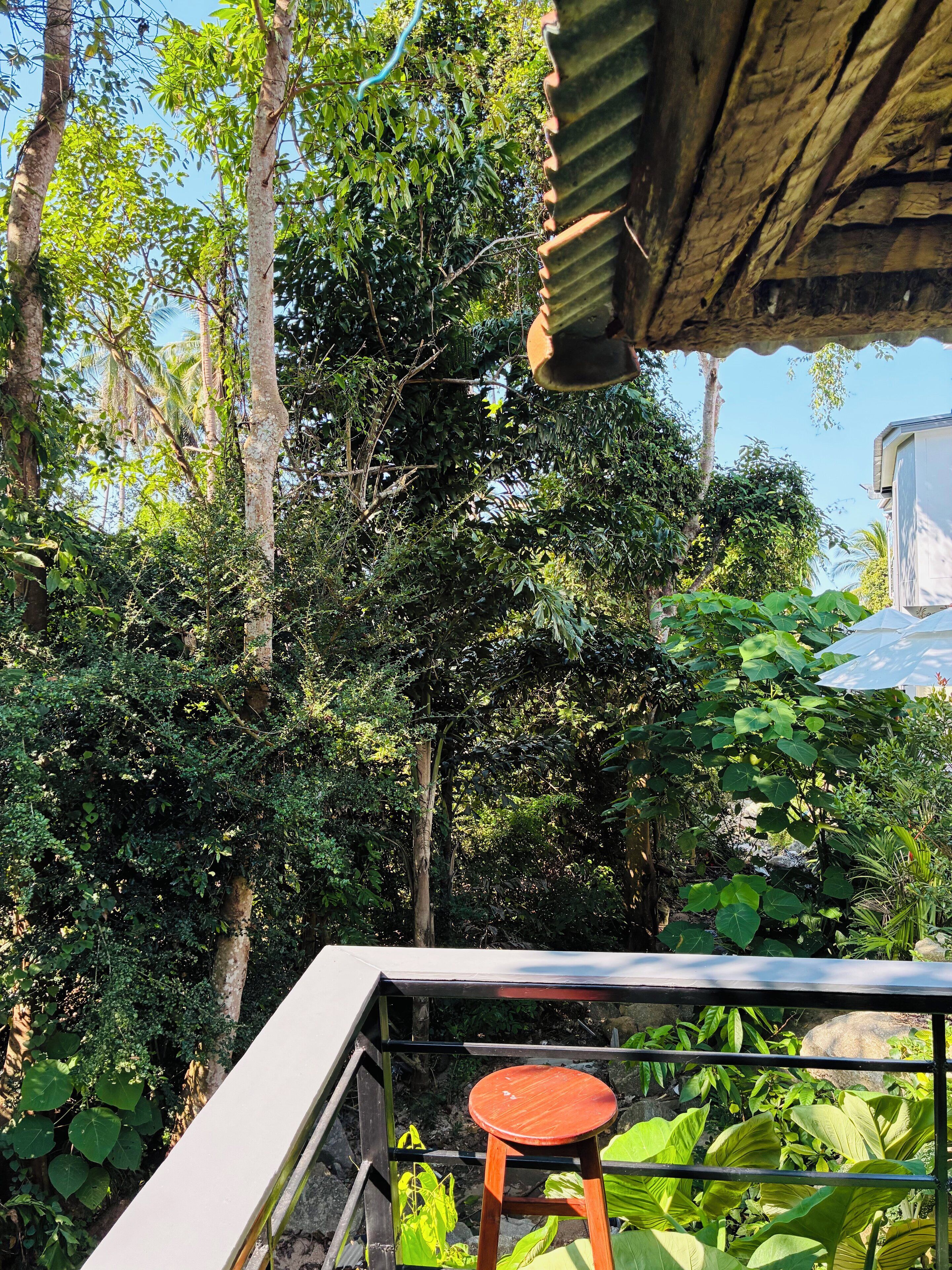 A balcony with a wooden stool, surrounded by lush greenery and a metal railing.