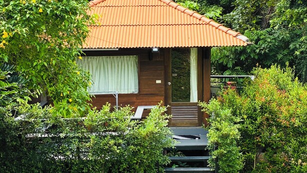 Een klein houten vakantiehuisje met pannendak, omringd door weelderige begroeiing en een tuin met een grindpad dat leidt naar een tafel en stoelen.