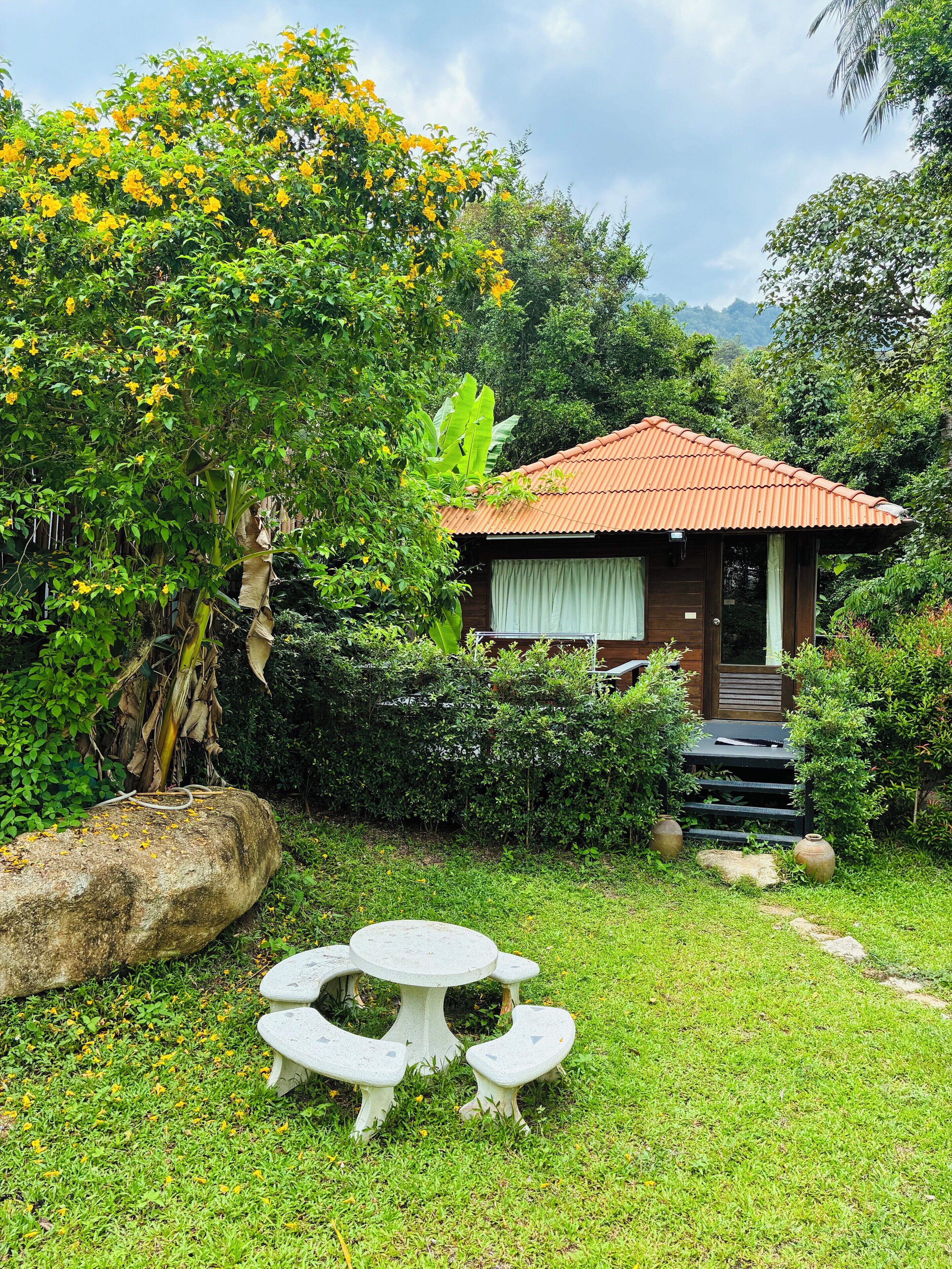 A small house with a red roof, surrounded by lush greenery and a stone table with benches in the foreground.
