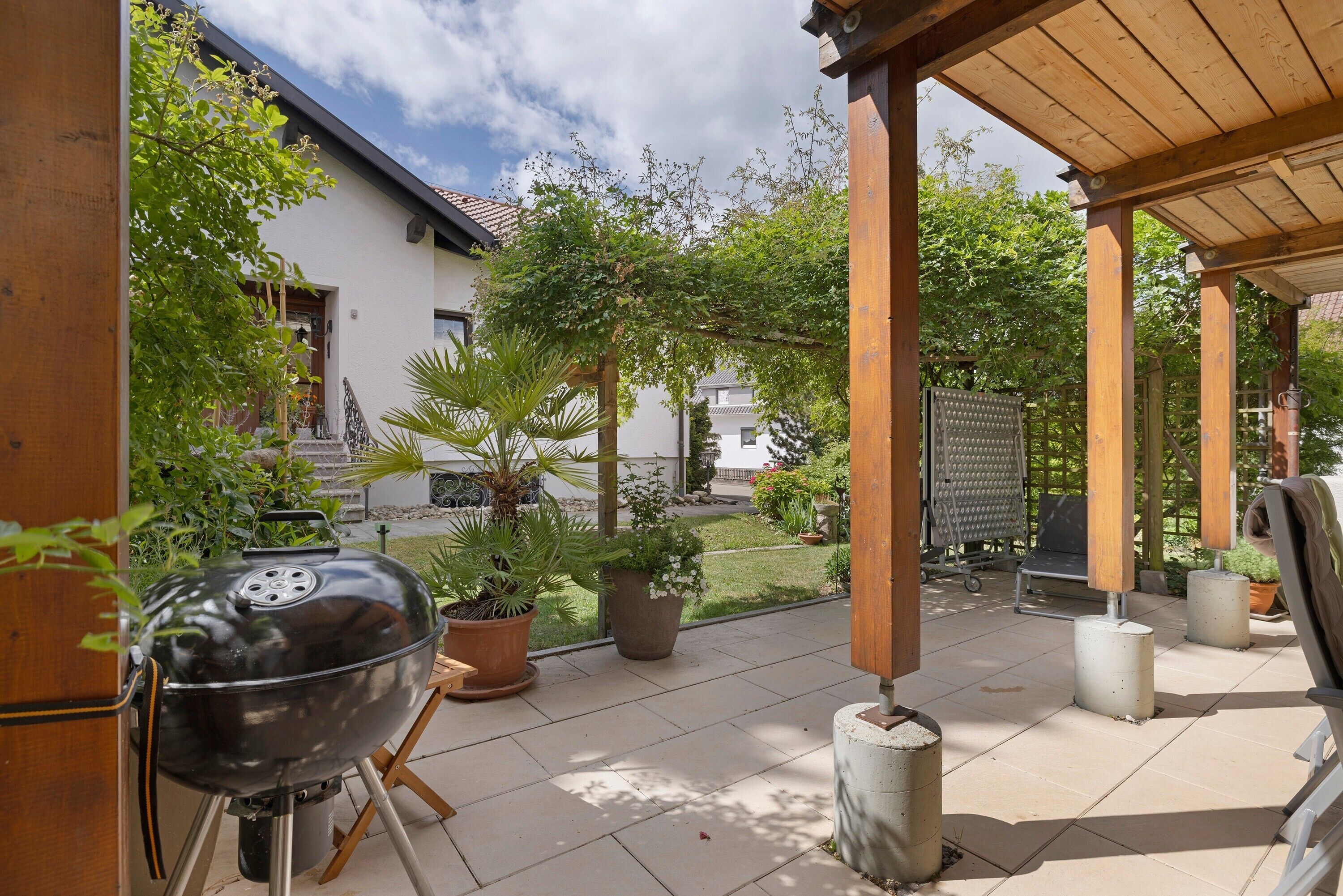 A covered patio area with a barbecue grill, potted plants, and a view of a house in the background.