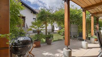 A covered patio area with a barbecue grill, potted plants, and a view of a house in the background.