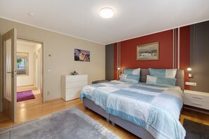 A bedroom with a bed, bedside tables, a nightstand, a dresser, a door, a hallway, and a painting on the wall.