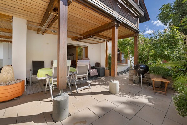 An outdoor patio area with a wooden pergola, a barbecue grill, a seating area with a table and chairs, and a potted plant.