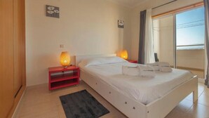 A bedroom with a bed, bedside lamps, a red nightstand, a black rug, and a window with curtains. - Charming 1-bedroom apartment in Portimão (Portimão)