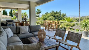 A covered outdoor seating area with a sofa, chairs, and a table, overlooking a lush landscape with palm trees and a view of the sea.