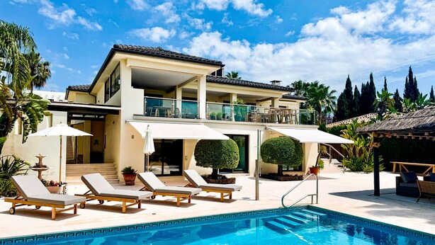 A two-story house with a swimming pool, lounge chairs, and umbrellas.