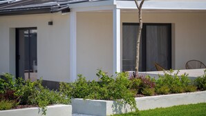 A modern building with a flat roof, a door, and a window. There is a small tree and a garden area in the foreground. - Kymè - One Bedroom Apartment, Sleeps 4 (NOTO)