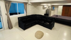 A modern living room with a black sectional sofa, a small round rug, and a kitchen area with a microwave and stove. - Cozy Apartment GM8 (Koh Samui)