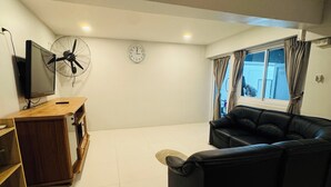 A living room with a black leather sofa, a wooden TV stand, a wall-mounted fan, and a window with curtains. - Cozy Apartment GM8 (Koh Samui)