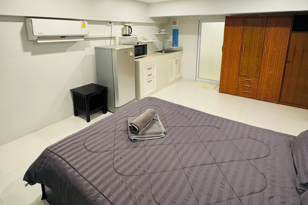 A modern bedroom with a large bed, a small kitchenette, a wardrobe, and an air conditioning unit.