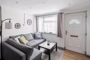 A modern living room with a grey sofa, a small table, a window with curtains, and a white door. - Cosy Central Home with Private Garden & Fast Wifi, Comfy 3BR Home • Contractor Friendly • Town Centr... (Colchester)