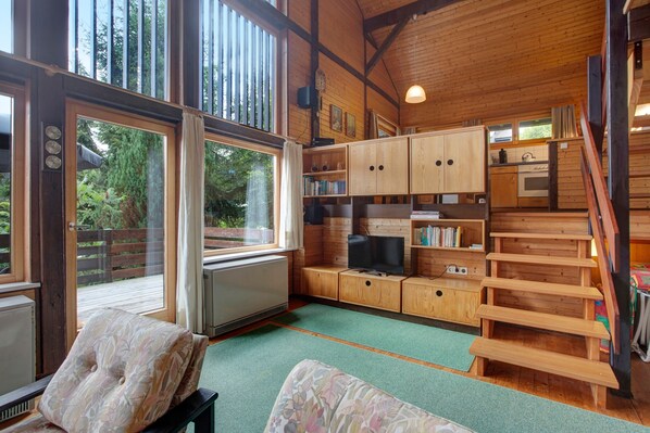 A wooden cabin interior with a television, bookshelves, a staircase, and a view of the outdoors.