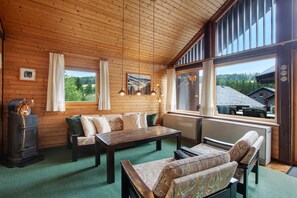 A cozy cabin interior with wooden walls, a sofa, a table, and a large window offering a view of a forested area.