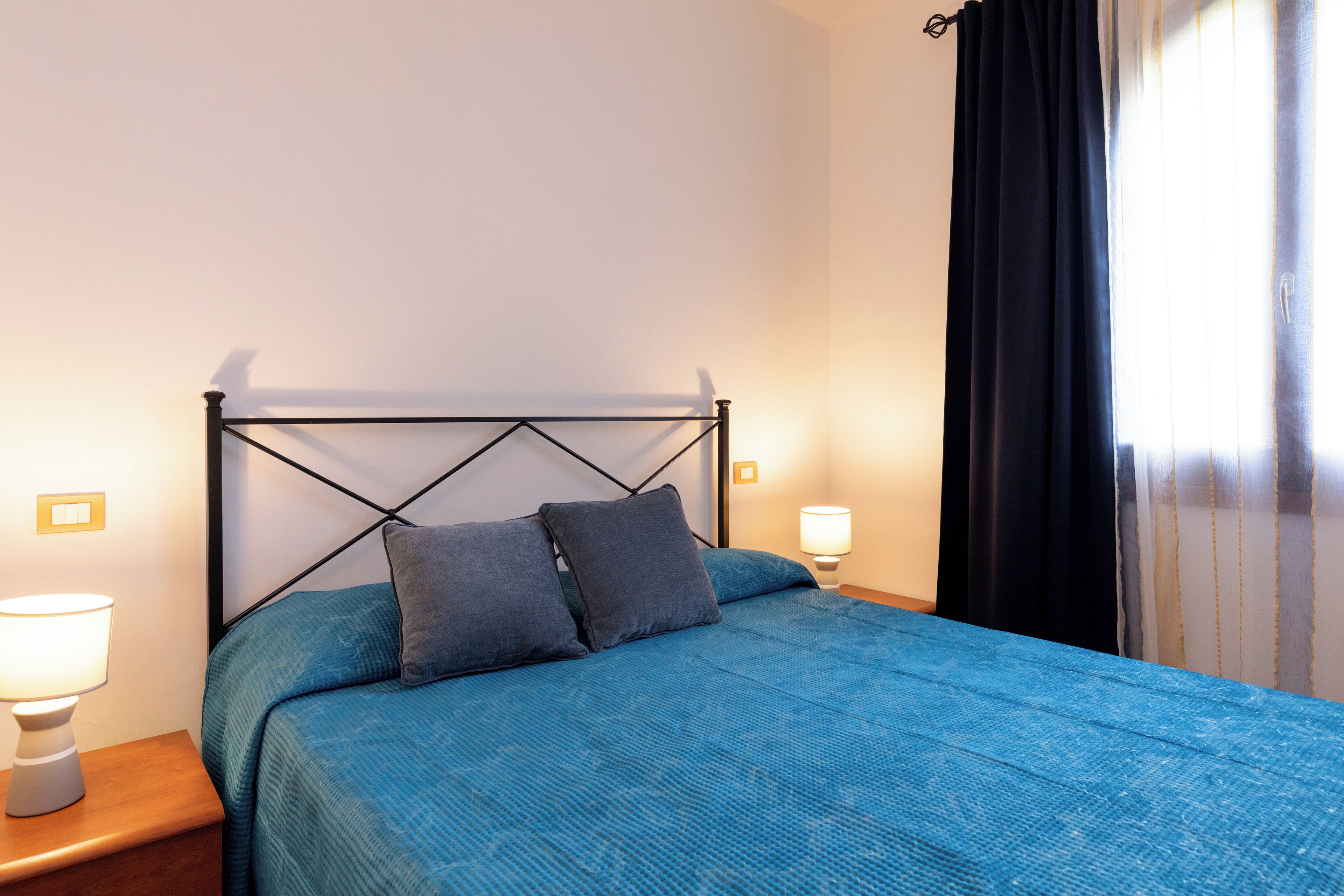 A bed with a blue bedspread and two pillows, a bedside lamp, and a window with black curtains.